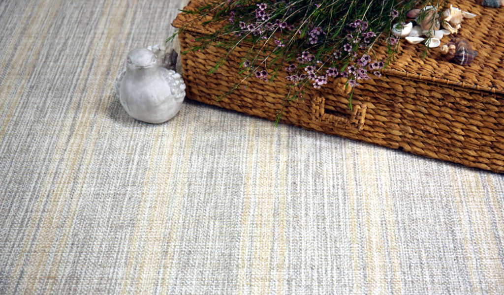 Linen Band, Color: Silver/ Sand 23/5702 .Image shows Linen Band carpet and props - wicker basket, flowers, shells and miniature bird decor. Linen Band has stripes that runs down the length of the carpet. The linear design is created by 3 colors - silver, sand and white.