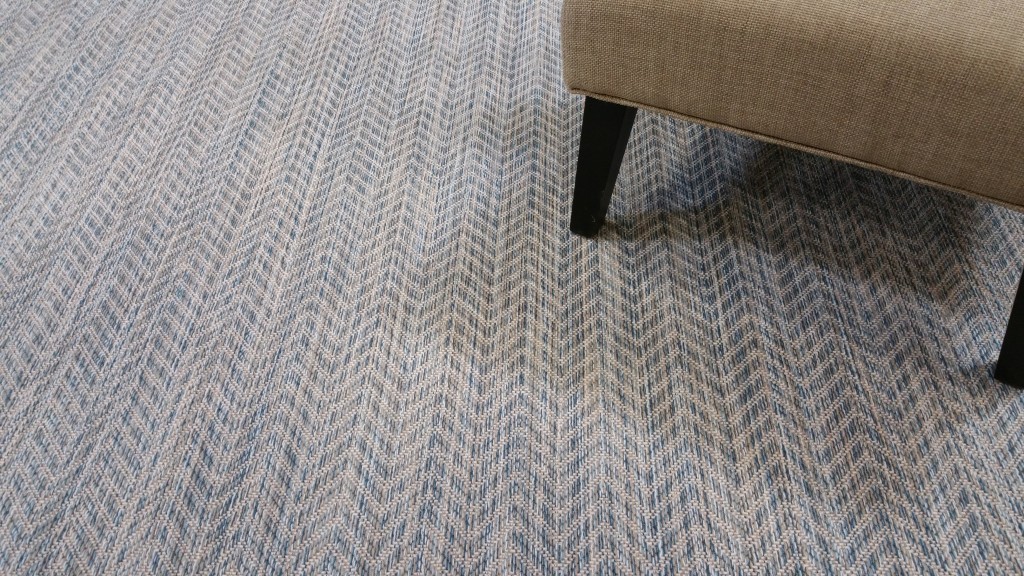Riverbed, Color: Shallow Water 41/5730 with chair as prop. Riverbed is a herringbone pattern created with striated yarns of blue, camel and beige. Image shows several pattern repeats of the carpet.
