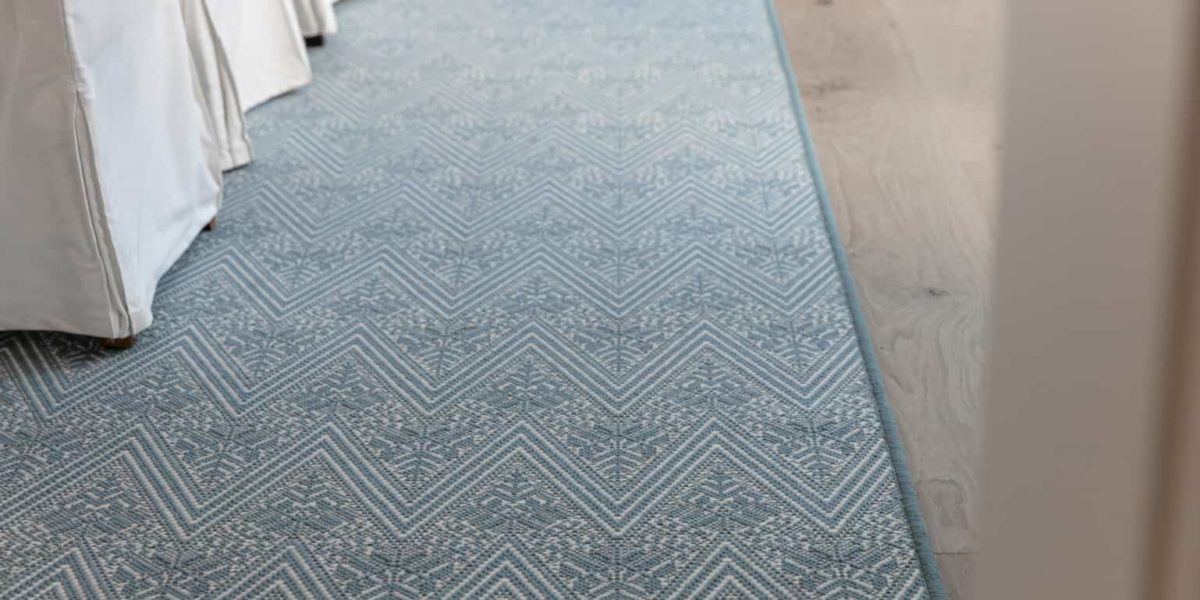 Fleur, color: Lt Denim 17/5742 rug showing serged edges and chairs. Fleur is fabricated with 2 yarn colors. denim and white that create an intricate flame stitch/ floral motif design. Image courtesy of Properties and The Beaubelle Group, both located in Orinda, CA
